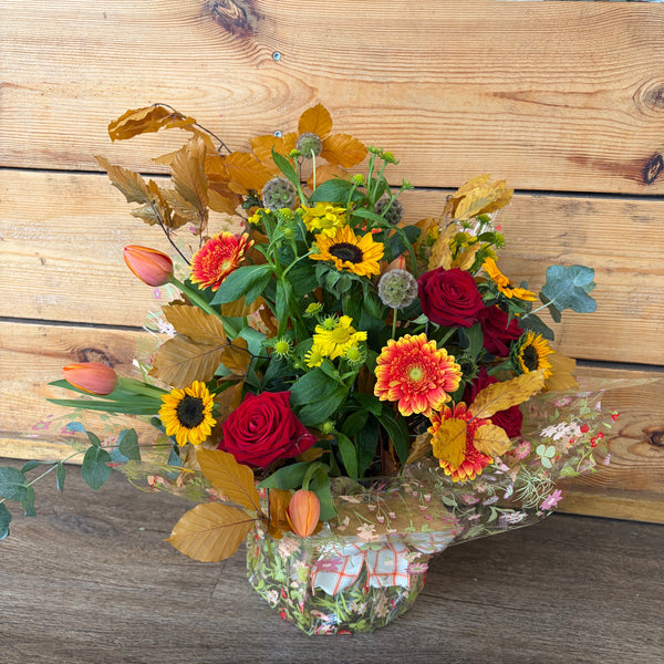 An autumn-themed floral arrangement in a clear glass vase, featuring red roses, yellow sunflowers, orange zinnias, and fall leaves, set against a rustic wooden plank background.