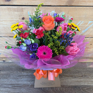A vibrant mixed flower bouquet in a clear plastic wrap with a bright orange ribbon and a small card at the base, set against a rustic wooden plank background.