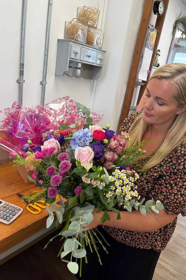 A woman with blonde hair wearing a black and orange floral patterned top holds a large, vibrant flower bouquet in a shop setting. The bouquet features pink, purple, blue, and white flowers with green foliage, including eucalyptus.