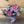 Load image into Gallery viewer, A vibrant floral bouquet featuring pink lilies, pink roses, pink gerbera daisies, and purple filler flowers, wrapped in clear cellophane with a pink ribbon, set against a rustic wooden plank background.
