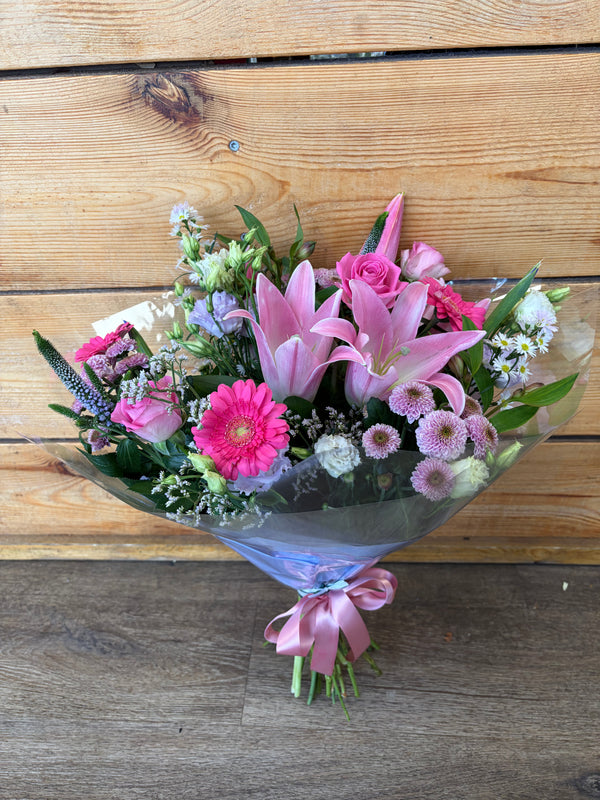 A vibrant floral bouquet featuring pink lilies, pink roses, pink gerbera daisies, and purple filler flowers, wrapped in clear cellophane with a pink ribbon, set against a rustic wooden plank background.