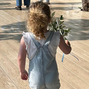 A toddler viewed from behind walking on a wooden boardwalk, wearing a light blue denim overall romper over a white sleeveless top and holding a small green leafy sprig.