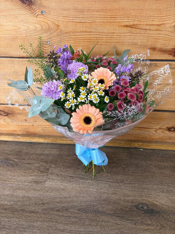 A vibrant mixed flower bouquet wrapped in clear cellophane and tied with a light blue ribbon, set against a rustic wooden plank background.
