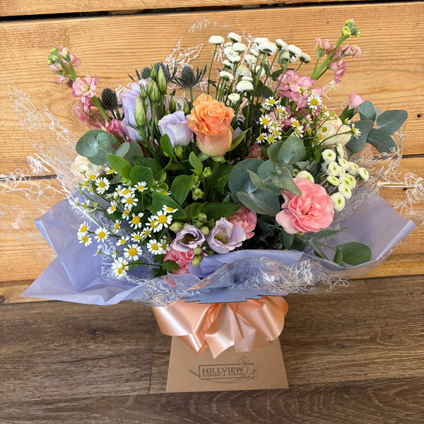 A mixed floral bouquet with pink roses, peach roses, white daisies, lavender, and green foliage, wrapped in pale blue paper and a peach ribbon, with a small card reading 'HILFIGUR' at the base, set against a wooden plank background.